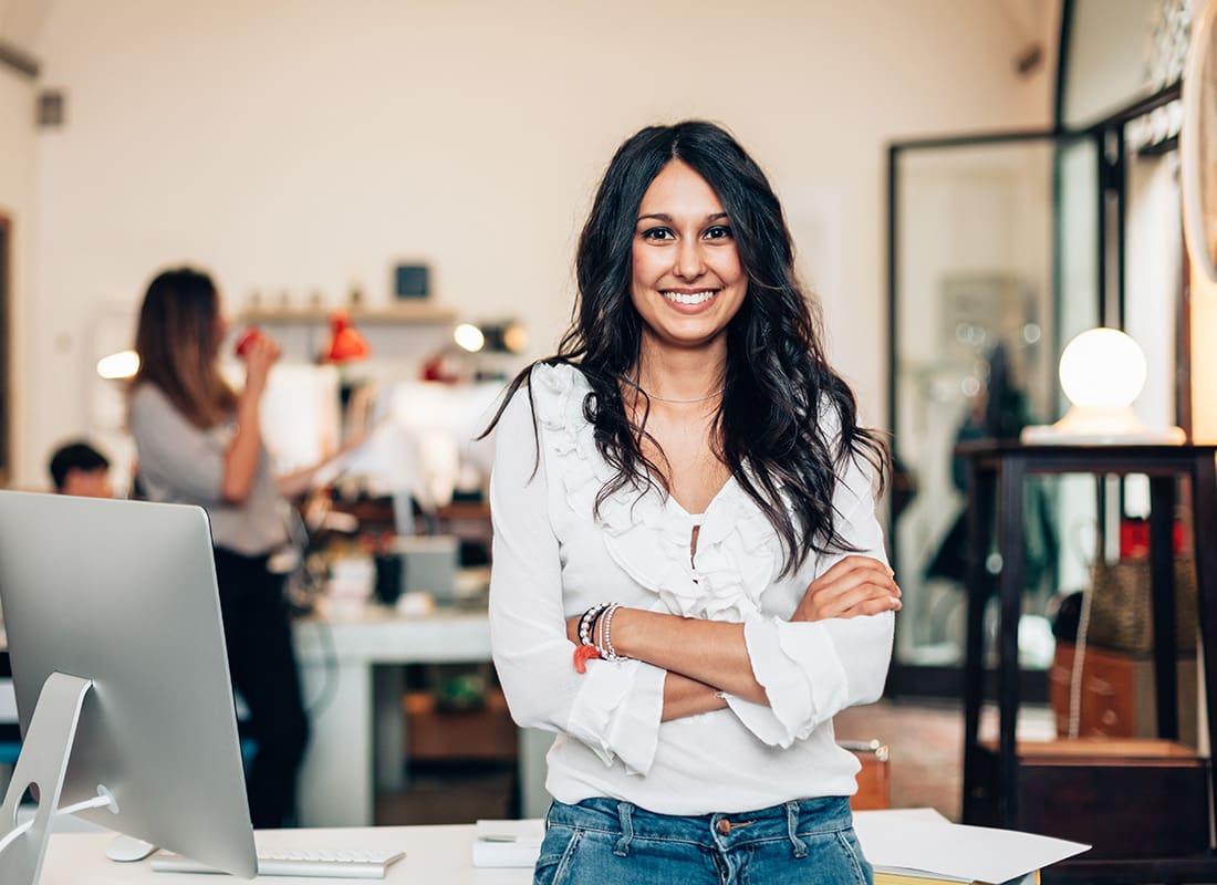 Business Owners Insurance - Portrait of a Cheerful Young Business Woman Standing Next to a Desk with a Monitor in a Small Office