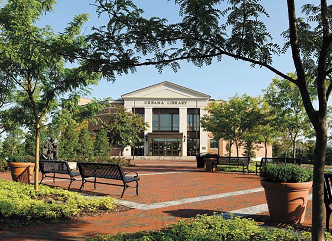 Read Our Reviews - Urbana Library Building with a Paved Courtyard in the Front with Benches and Green Trees on a Sunny Day